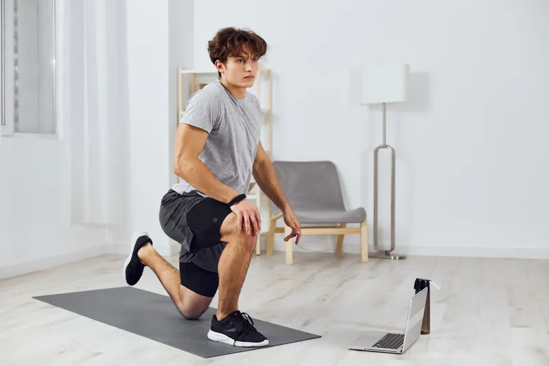 Мужчина в серой комнате занимается спортом с гантелями, следуя онлайн-уроку на часах