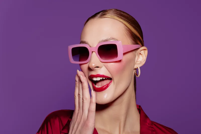 Stylish woman with pink sunglasses and red lipstick smiles with hand near mouth on purple background. Concept of fashion, beauty, glamour, and confident female model.