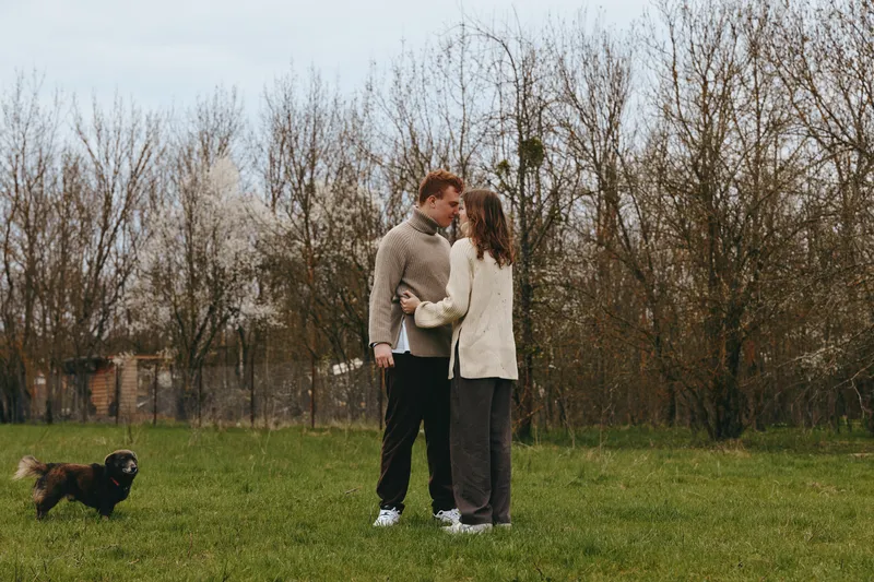 Two people embrace in a calm park as a small dog runs toward them, autumn trees in the background. A candid lifestyle moment highlighting connection, companionship, and everyday affection in bright