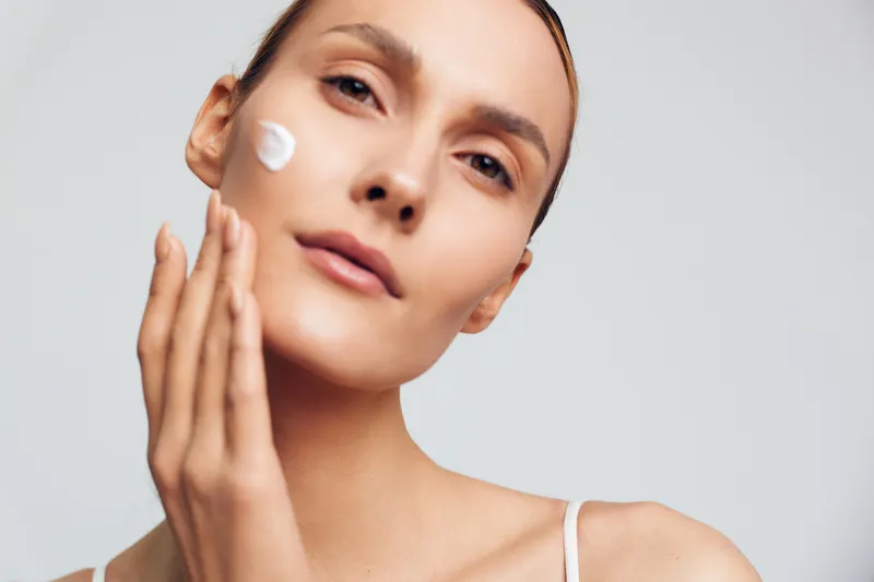 Young woman applies moisturizing cream on face with fingers closeup natural skin care beauty treatment isolated on light background with soft expression and clean complexion.