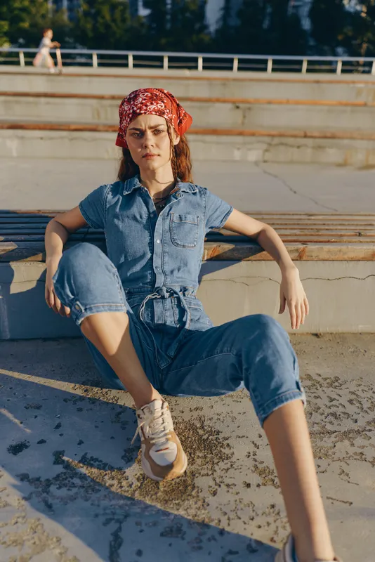 lifestyle woman in modern boho-western denim street style, wearing a red bandana and casual sneakers, sitting outdoors in warm natural light, film color effect