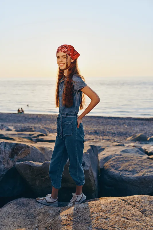 lifestyle woman in modern boho-western denim street style stands by the sea during golden hour with film color warmth and relaxed happy expression