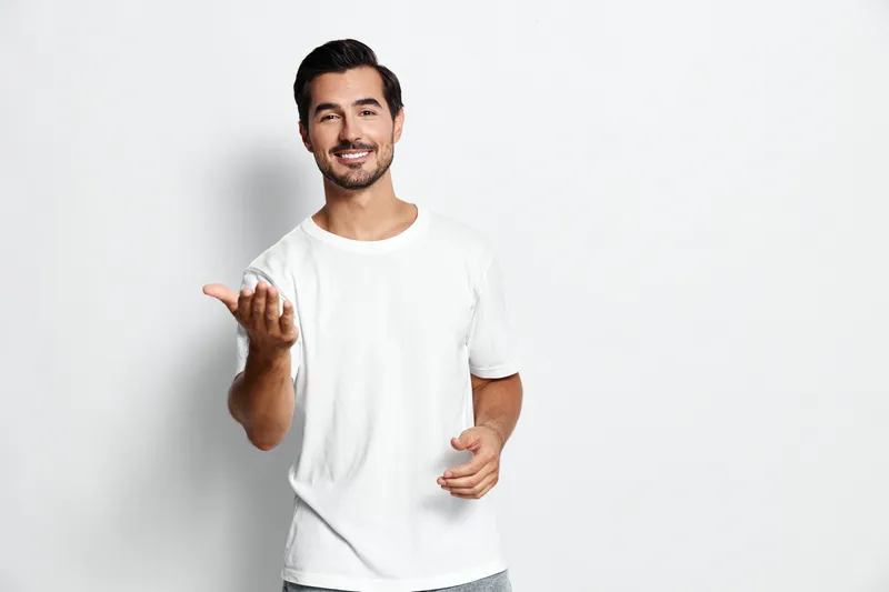 Full body smiling cheerful young man in casual white t-shirt looking at camera with hand gesture, isolated on plain solid gray background studio portrait. People lifestyle concept