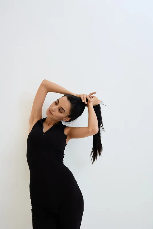 young woman stretching with arms raised wearing black sleeveless jumpsuit, isolated on white background, fitness and relaxation concept
