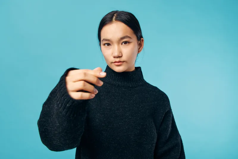 Young woman in black sweater posing with hand extended towards camera on blue background. Female model with serious expression and long dark hair in studio portrait.