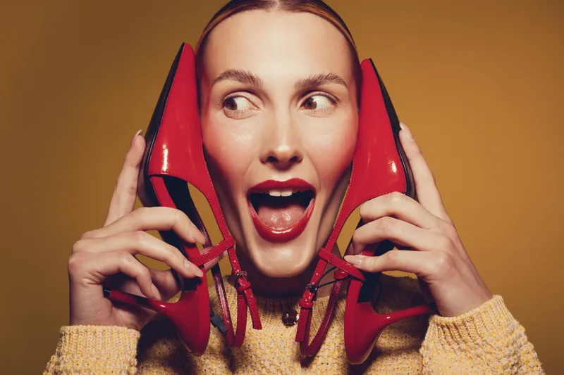 Young woman with red lipstick holds red high heel shoes near her face, smiling and looking sideways on yellow background. Concept of fashion, style, and playful mood.