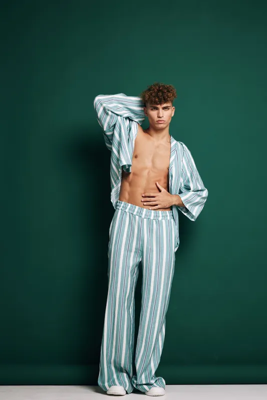 Young man with curly hair wearing striped pajamas posing with hand on head and stomach against green background in studio. Casual sleepwear and relaxed style.