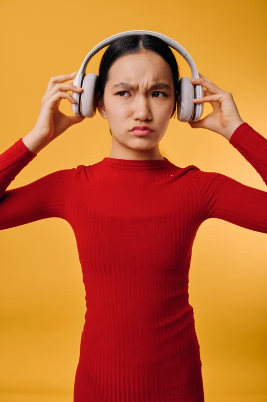 Young woman wearing headphones with confused expression adjusting wireless audio device on yellow background. Female person in red sweater showing doubt or discomfort while listening.
