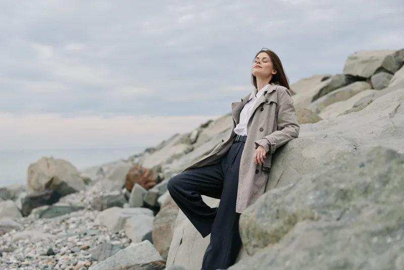 Young woman in a long coat leans on large rocks at a rocky beach under a cloudy sky. She looks relaxed and contemplative, enjoying nature and solitude outdoors.