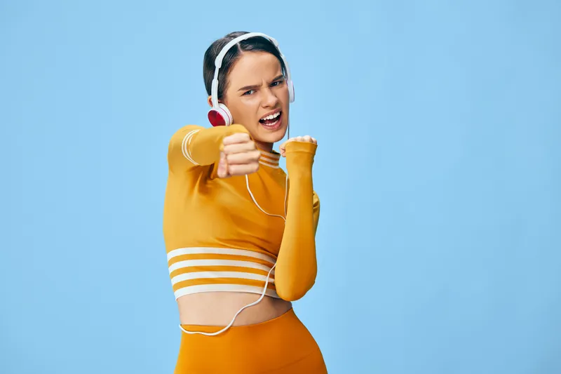 Athletic woman in orange workout set with headphones, striking a dynamic pose against a blue background, conveying energy, sport focus and active fitness.