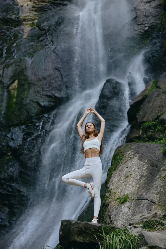 woman practicing yoga in nature, standing on one leg near waterfall, wearing white fitness clothes, serene outdoor landscape, calm and focused mood