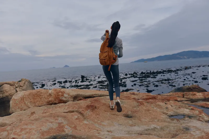 Young female traveler exploring rocky coastline, wearing a backpack, reflecting on nature s beauty, showcasing adventure and freedom