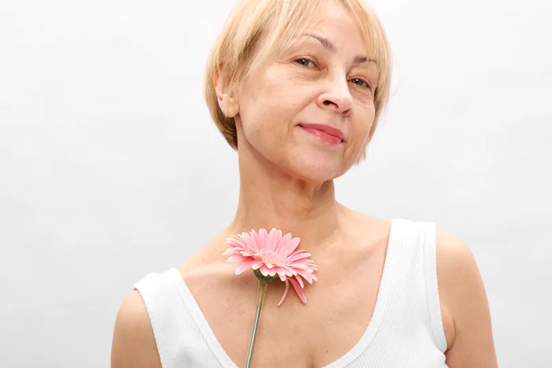 Portrait of a mature woman holding a pink gerbera flower, spring calm beauty with aging grace, minimalist white studio background and soft natural light