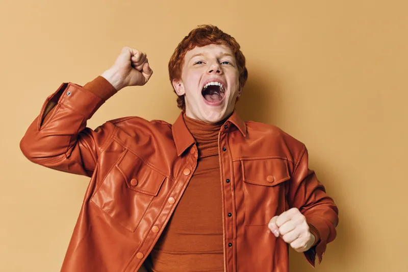 Young man with red hair wearing orange leather jacket and turtleneck celebrating with raised fist and open mouth on beige background.