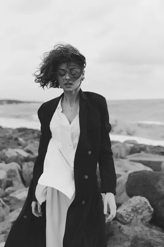 Monochrome fashion scene of a windblown model on a rugged coastline, wearing a long dark coat over a white shirt and light trousers, standing among rocks with a vast sea horizon