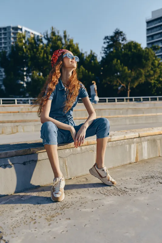 lifestyle woman in modern boho-western denim street style sits outdoors wearing sunglasses and a red bandana, captured with film color tone under natural sunlight