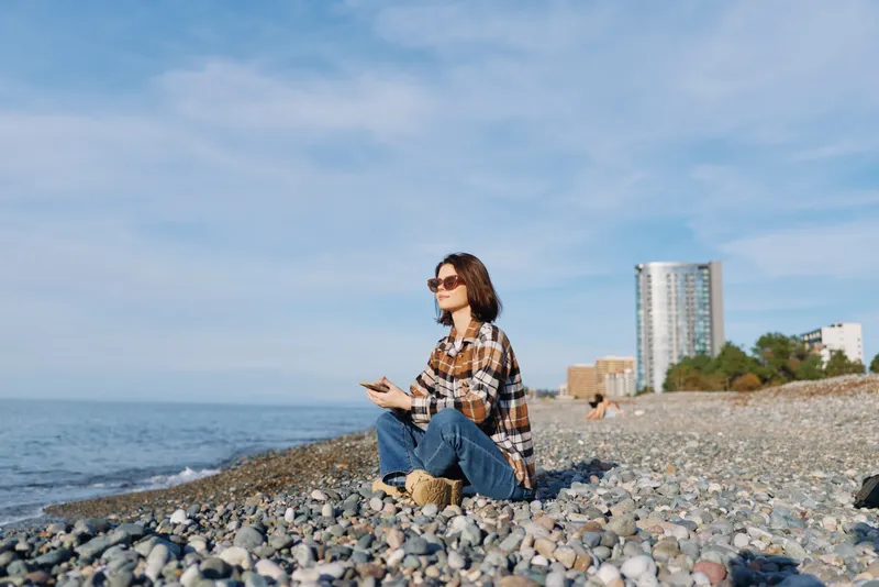 Woman sits on a pebble beach by the sea, wearing a plaid shirt and denim jeans, gazing toward the water under a clear sky with distant buildings.