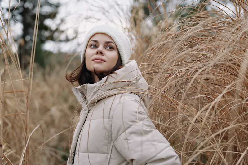 Woman in a warm beige jacket and white beanie stands among tall grass, posing for a moody outdoor portrait with soft natural light and a peaceful atmosphere.