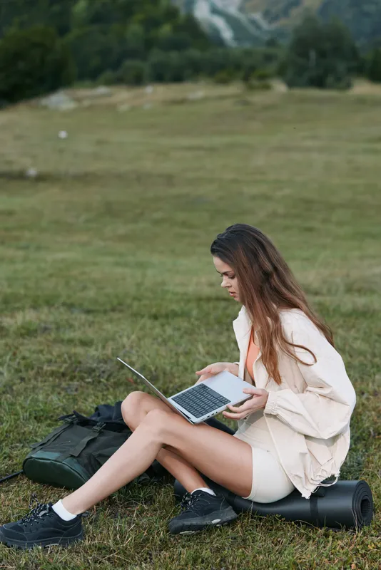 A woman sits on grass with a laptop in an open field, enjoying an outdoor workspace. She appears focused and calm, creating a productive and relaxed atmosphere.