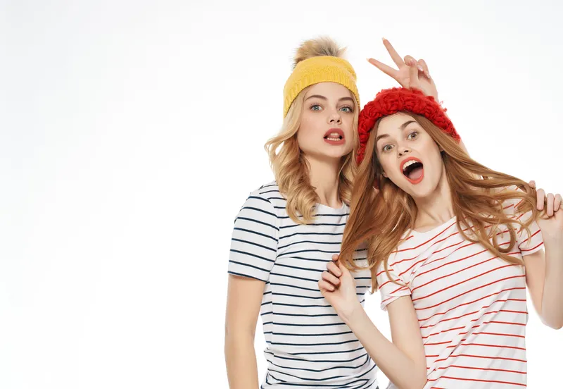 Две женщины в кепках обнимаются на светлом фоне, передавая дружеские эмоции