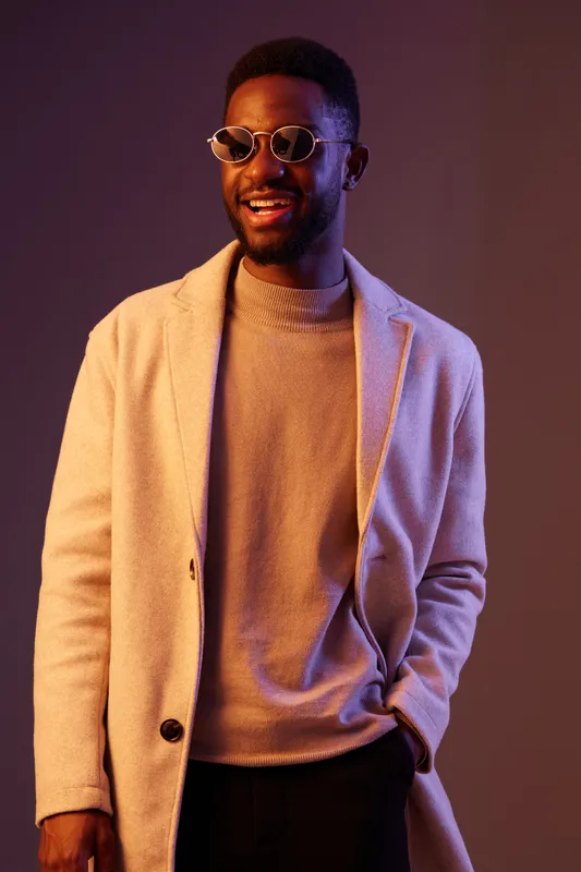 Stylish young man with sunglasses wearing a beige coat and a gray sweater, smiling against a colorful gradient background, embodying confidence and modern fashion