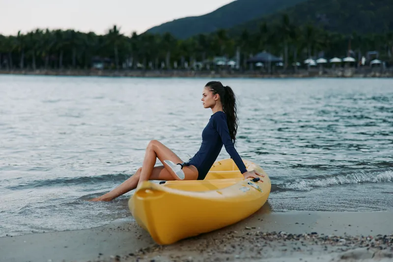 Woman sitting on yellow kayak at the beach, wearing swimwear, relaxing by calm sea during summer sunset with tropical palm trees in background.