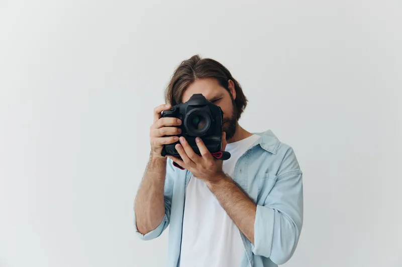 Мужчина-хипстер, фотограф, в студии на фоне белого фона смотрит в видоискатель камеры и делает снимки при естественном свете из окна