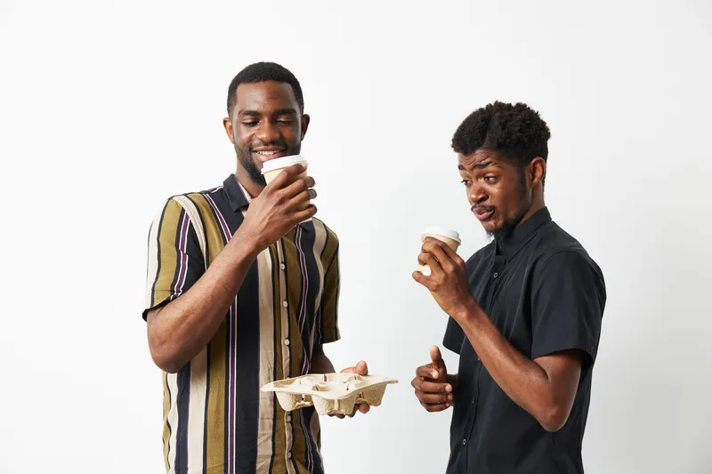 Two smiling African American men enjoy coffee together, showcasing friendship and joy. Their stylish outfits highlight a contemporary urban vibe.