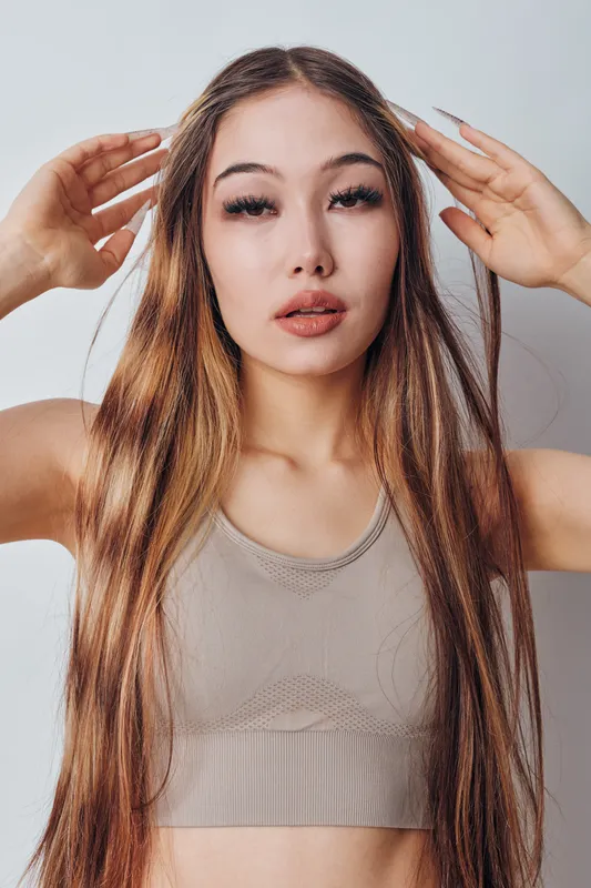 Young woman with long straight hair wearing sleeveless top posing with hands near head, looking at camera against light background in studio portrait. Beauty, fashion and confident model