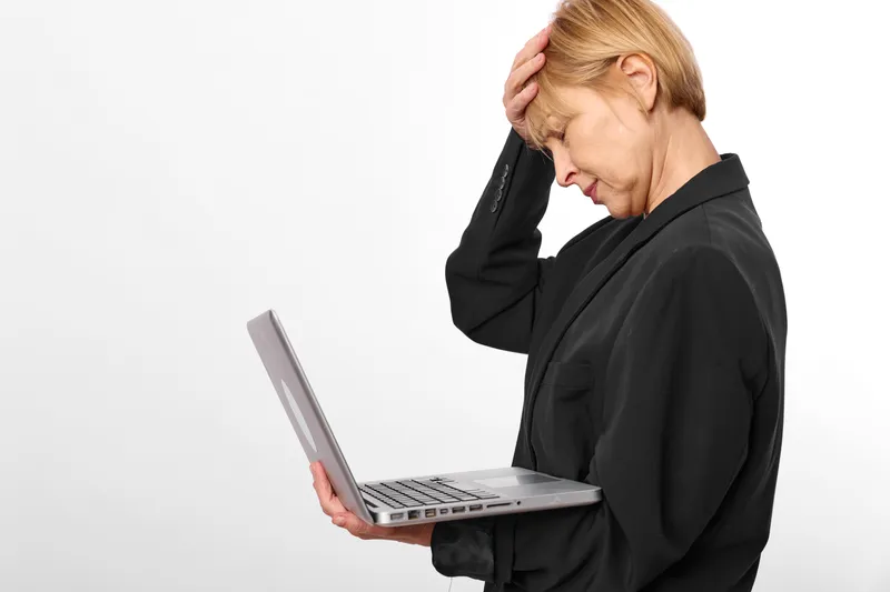 Stressed woman with blonde hair holds an open laptop, hand on her head, showing headache and work fatigue, concept for anxiety, burnout, mental health, office pressure