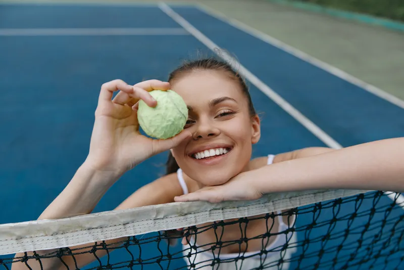 Young female tennis player smiling with a tennis ball on a court, showcasing sporty attire and vibrant energy on a sunny day
