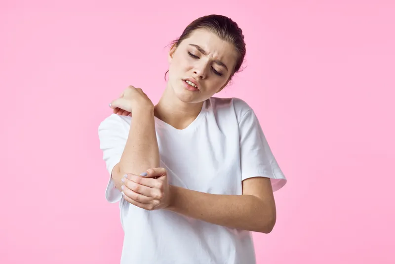 Женщина в белой футболке держится за шею, испытывая боль, на розовом фоне