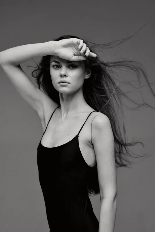 Black and white portrait of a woman with long hair posing with her arm raised, showcasing elegance and grace.