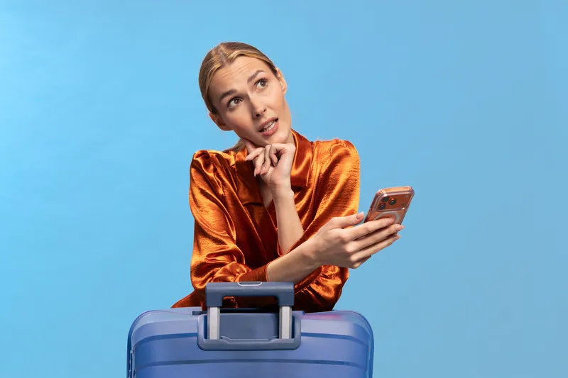 Thoughtful young woman in orange satin blouse leaning on blue suitcase holds smartphone and looks up. Concept for travel planning, waiting, and mobile communication with blue background.