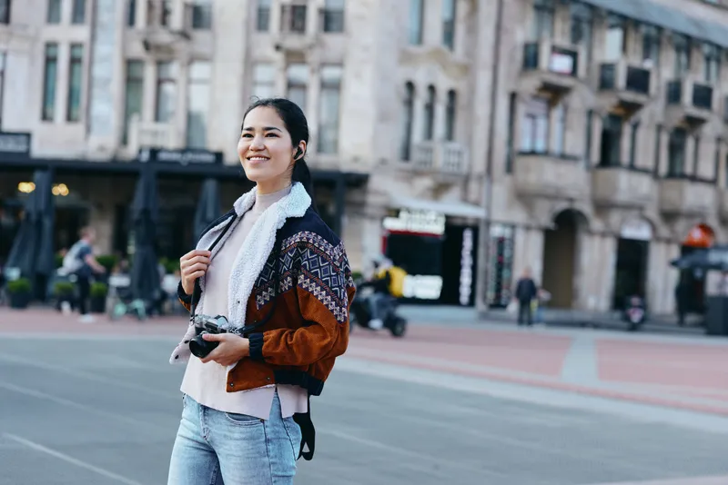Woman photographer on a city street with a camera, wearing a patterned jacket and scarf, smiling while exploring urban architecture and outdoor travel moments.