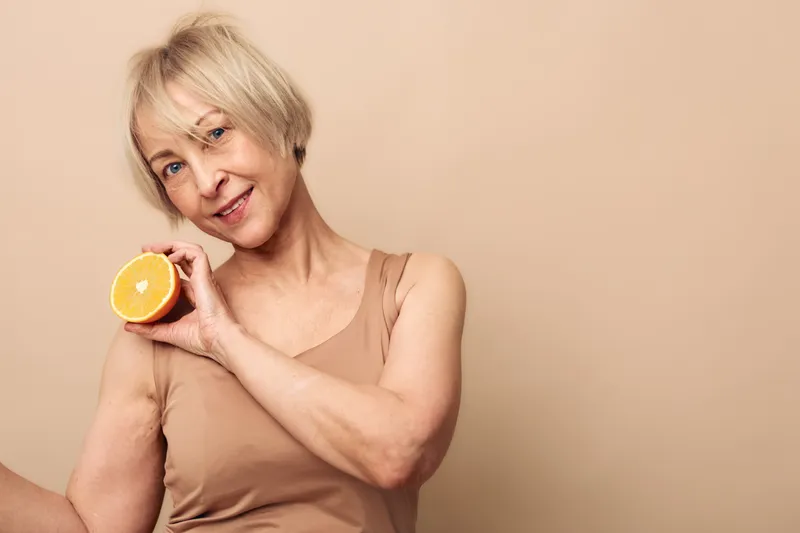 Smiling mature woman with short blonde hair holds fresh orange slice near shoulder on beige background. Concept for healthy lifestyle, nutrition, wellness and natural beauty in middle age.