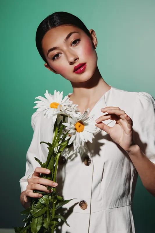 Elegant young woman with smooth makeup holding fresh chamomile flowers. Close-up on pastel background. Concept of beauty, creative fashion, natural elegance, summer mood.