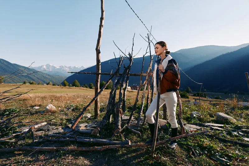 Woman in hiking jacket standing by rustic fence in mountain meadow, enjoying nature and rural countryside with distant mountains, calm outdoor lifestyle and exploration scene.