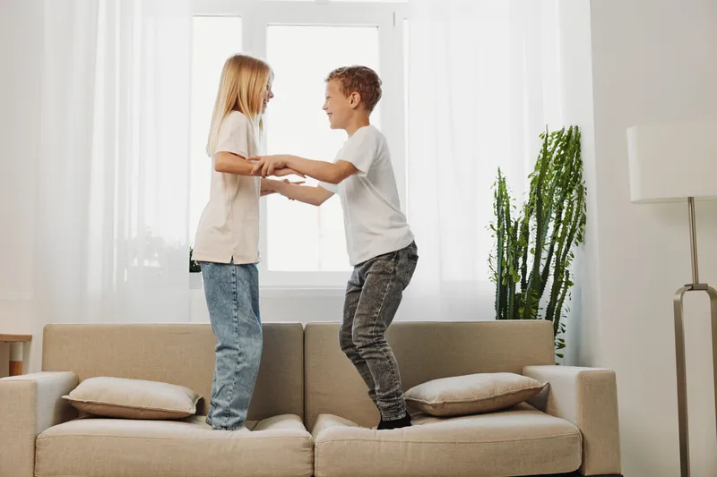 Two children standing on a sofa holding hands and smiling in a bright living room with natural light and modern decor.