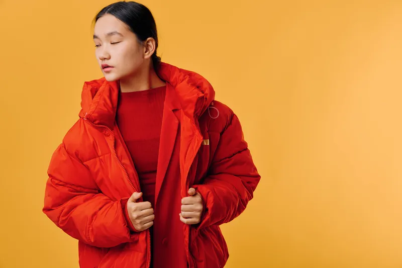 Woman wearing warm red puffer jacket and sweater standing against yellow background with closed eyes, fashion style, winter clothing, studio portrait with calm expression.