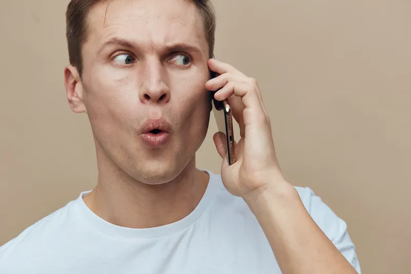 Confused man talking on phone with surprised expression, looking aside against plain neutral background, casual white t-shirt, indoor portrait. People lifestyle concept, emotional reaction.