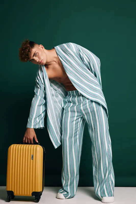 Young man in striped casual outfit holding yellow suitcase standing on white floor with dark green background, fashion and travel concept with minimalism and relaxed posture.