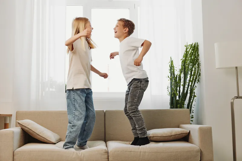 Two happy children jumping on a beige sofa in a bright living room with a tall green plant near the window. Concept of childhood joy, playfulness, and family comfort.