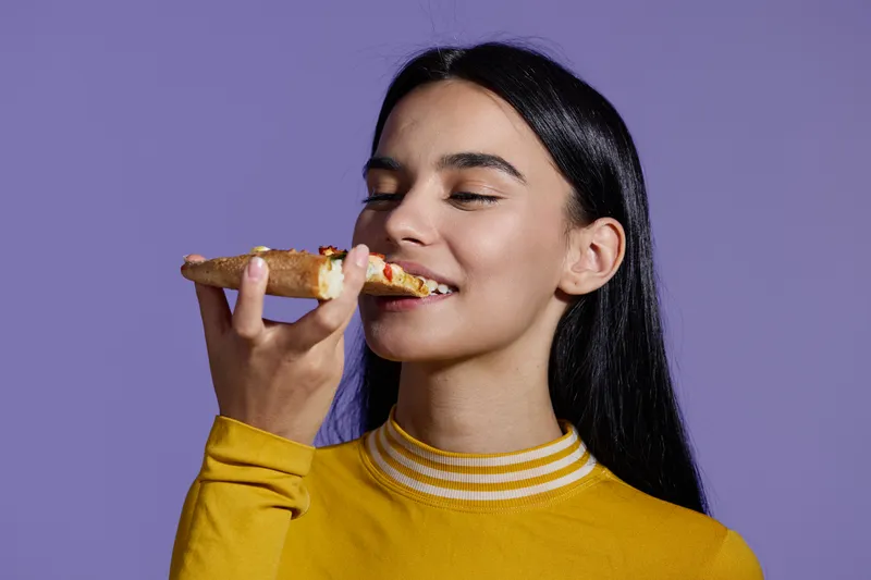 Sport fitness woman enjoying a slice of pizza, wearing a yellow top against a purple background, showing balance between healthy lifestyle and indulgence.