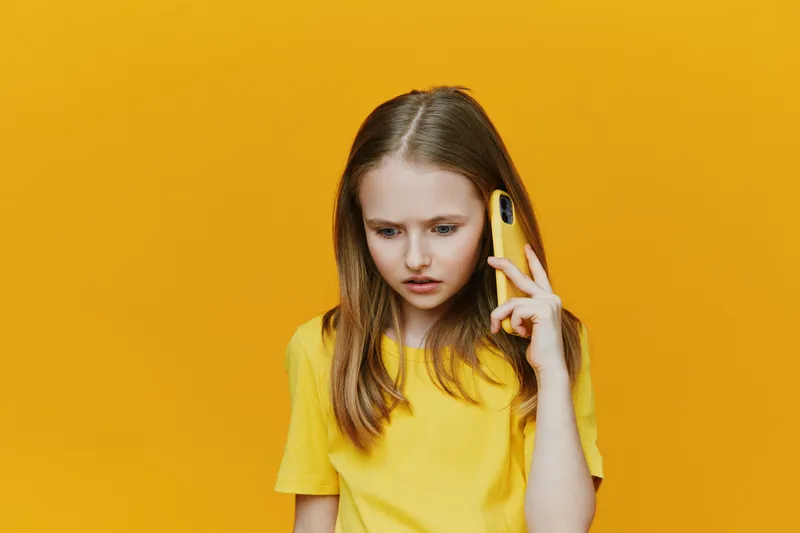 Young girl with long brown hair in a yellow shirt looking concerned while holding a yellow phone against a vibrant yellow background, expressing emotions of worry and anxiety