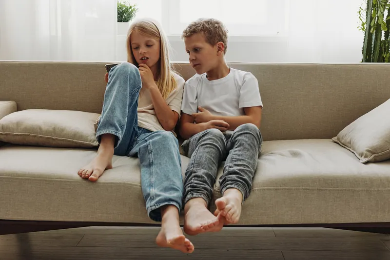 Two children sitting barefoot on a beige sofa, focused on a smartphone screen, casual clothes, bright natural light from window behind. Concept for childhood, technology use.