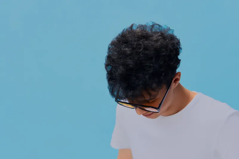 young man with curly hair wearing glasses and white t-shirt looking down against bright blue background, minimalistic style, casual young adult portrait