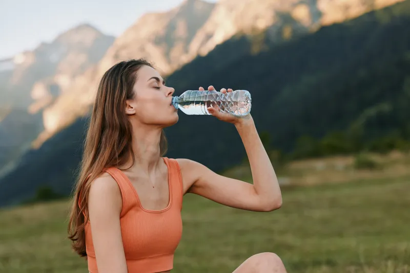 An athletic young woman drinks from a bottle while enjoying a scenic mountain landscape, wearing a sleeveless top, exuding health, energy, and outdoor vitality.