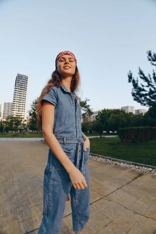 Lifestyle woman in modern boho-western denim street style wearing a red bandana outdoors with urban buildings and green trees in film color tones.
