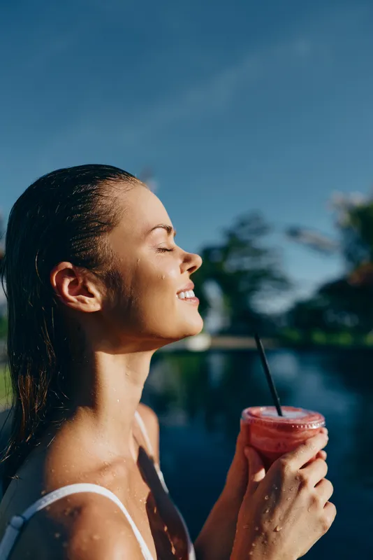 Sunny poolside moment features a smiling woman enjoying a pink smoothie, sipping through a straw, with water, palm trees and bright daylight in the background.